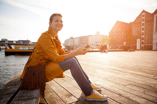 Woman wearing orange coat sitting on bench near canal of Copenhagen city and enjoying sun. Holding smart phone. Sunset, evening. Wooden floor, buildings in background. Nyhavn , Copenhagen, Denmark.