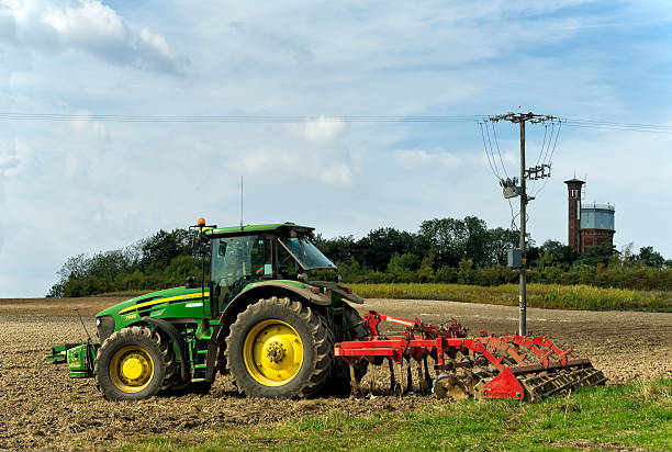 Sandringham, Norfolk, England - September 23, 2014: A tractor driving waiting in the cab of his John Deere tractor at the edge of a ploughed field near Sandringham, Norfolk, in Eastern England. Attached to the tractor is a Kongskilde cultivator. To the right is the Victorian water tower which supplies Sandringham House, the Queen’s Norfolk home.