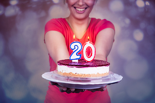 Beautiful young girl celebrating 20th Birthday, holding birthday cake with flaming candles in shape of 20. Purple, bokeh background