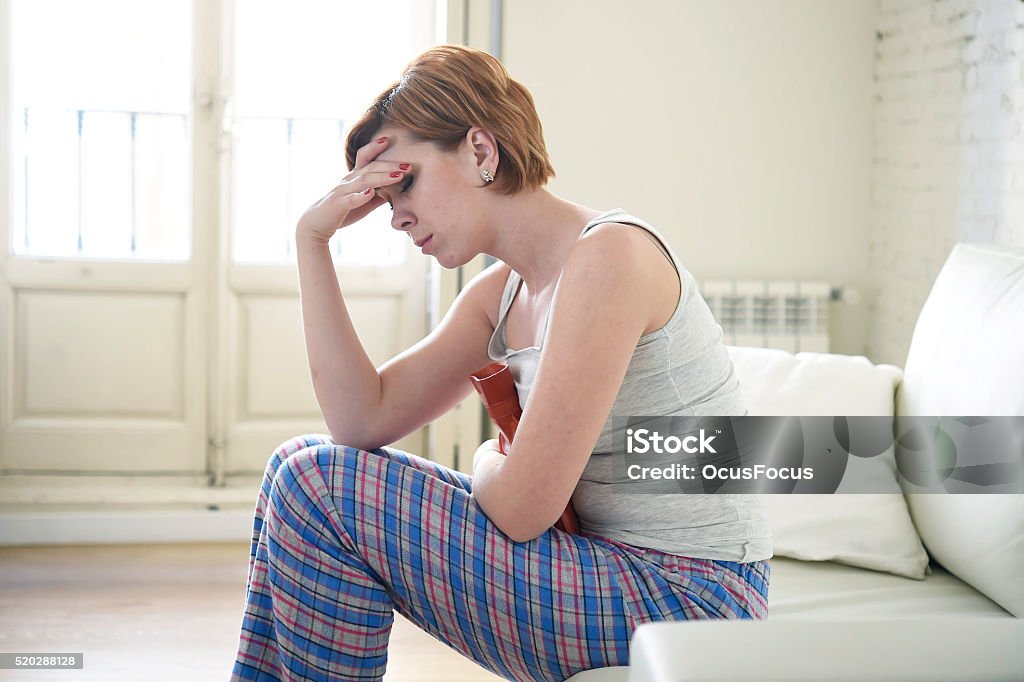 young woman holding hot water bottle suffering belly period pain young beautiful red hair woman holding hot water bottle in a hurting belly suffering stomach cramp and period pain sitting on home couch in painful face expression female menstruation concept Abdomen Stock Photo