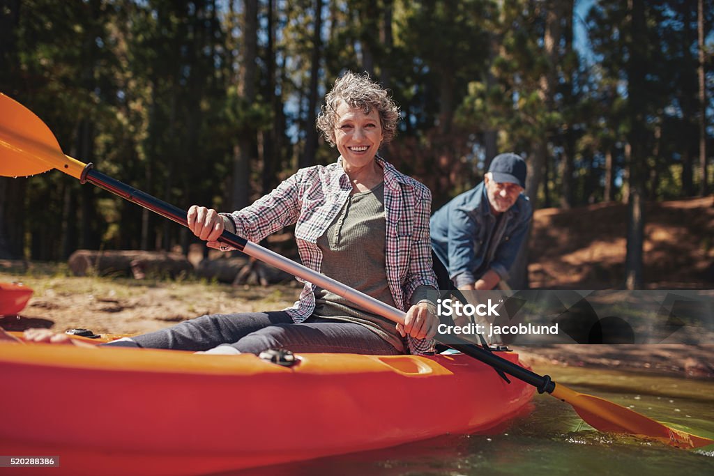 Happy senior woman in a kayak at the lake Portrait of happy senior woman in a kayak holding paddles. Woman canoeing with man in background at the lake. Kayak Stock Photo