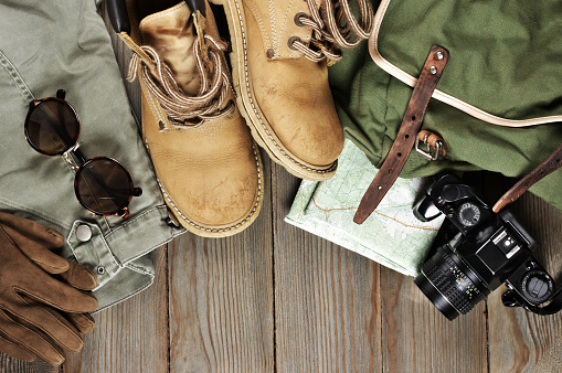 Travel accessories set on wooden background: old hiking leather boots, pants, backpack, map, gloves, vintage film camera and sunglasses. Top view point.
