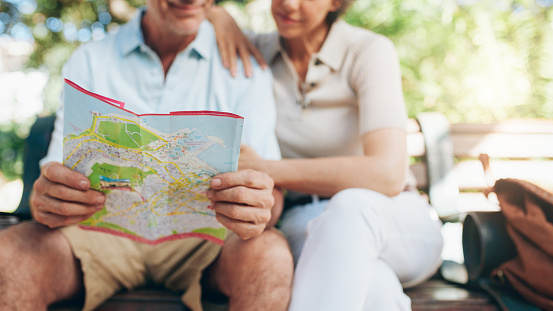 Cropped shot of couple of tourist sitting on a park bench using city map. Senior man and woman reading a map for direction. Focus on map.