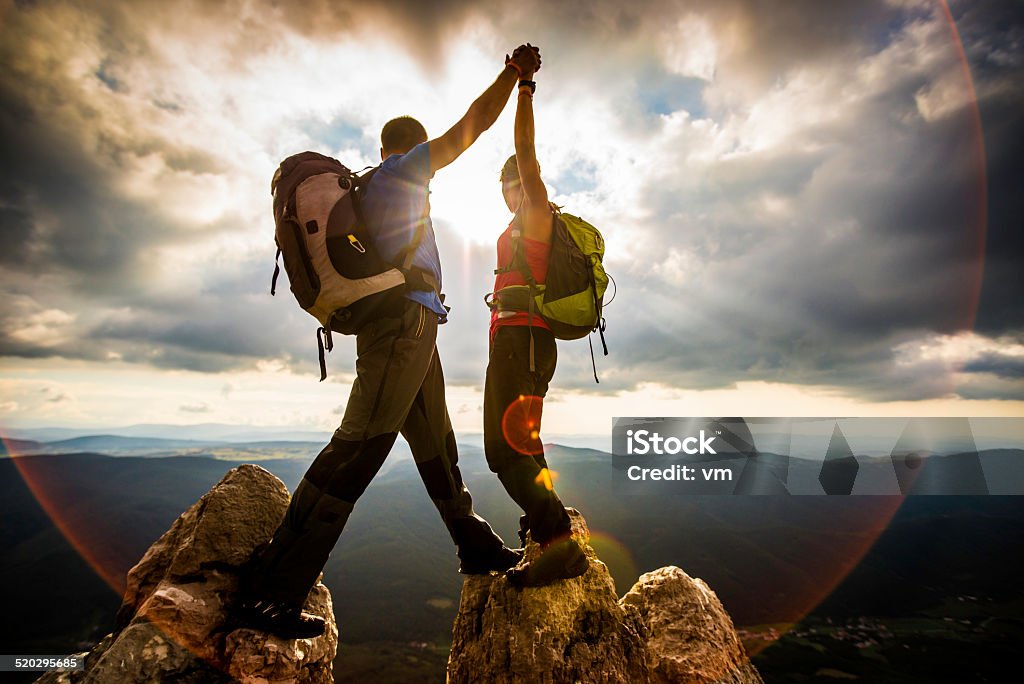 Couple on Top of a Mountain Shaking Raised Hands Couple hiker on top of mountain looking at beautiful sunset landscape, lens flare. Mountain Climbing Stock Photo