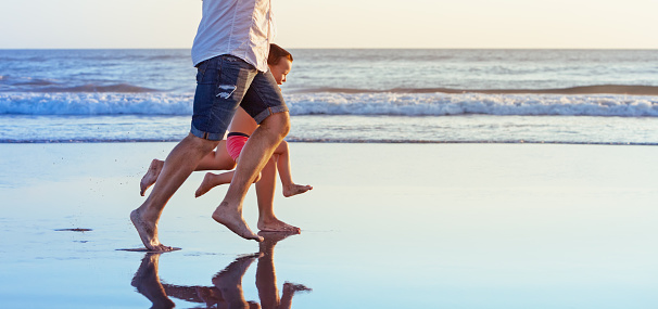 Barefoot legs in action. Happy family fun - parents with baby son running along edge of sea beach surf with sunset light. Active travel lifestyle, water activity and game on summer vacation with child