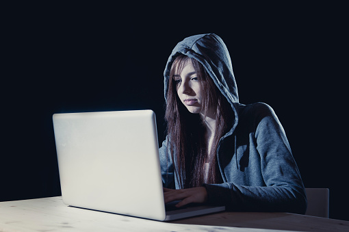 young attractive teen woman wearing hood on looking dark and dangerous hacking laptop computer system on black background in cybercrime or cyber crime and internet criminal concept
