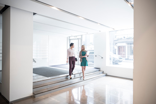 Two female professionals in modern foyer walking to office. Businesswomen talking on way to work in bright naturally lit modern office interior.