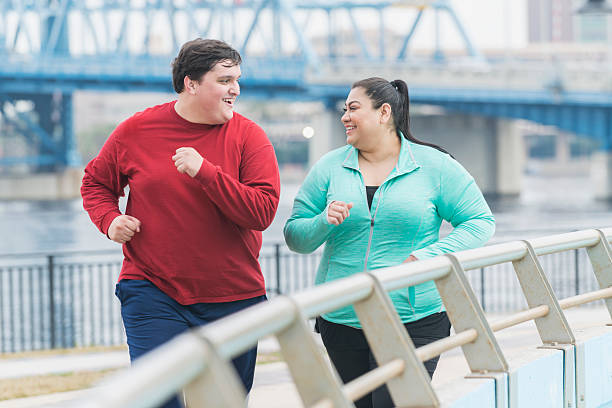 Overweight man and woman jogging in the city An overweight Hispanic woman and a young mixed race Hispanic and Caucasian man exercising together outdoors in an urban setting, running or jogging. They are smiling, looking at each other as they exercise. overweight stock pictures, royalty-free photos & images