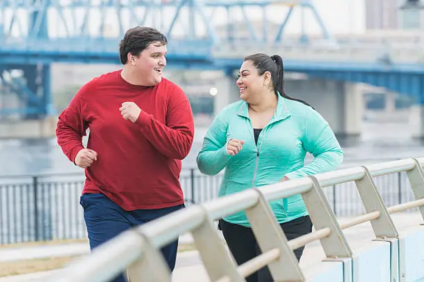 An overweight Hispanic woman and a young mixed race Hispanic and Caucasian man exercising together outdoors in an urban setting, running or jogging. They are smiling, looking at each other as they exercise.