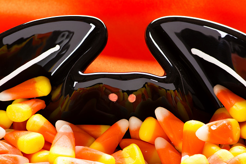 Close-up of a black bowl shaped like a bat filled with candy corn as a Halloween decoration and a tasty treat. Orange silk background.