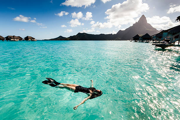Young Woman Floating in Wonderful Sea of Bora Bora A DSRL photo of a beautiful caucasian young woman floating in clear sea in Bora Bora, French Polynesia. She is wearing a black swimsuit and black fins. The day is sunny with scattered white clouds in the blue sky. At he background is mountain Otemanu, main natural landmark in Bora Bora. bora bora mountain stock pictures, royalty-free photos & images
