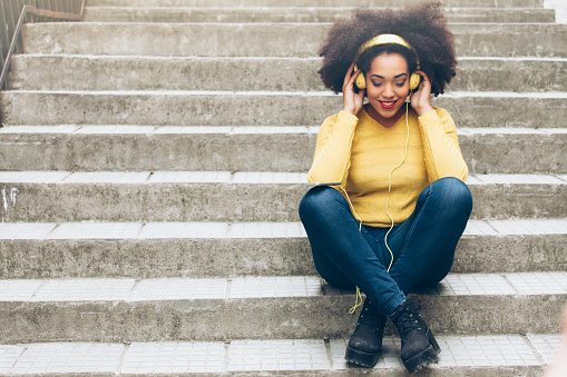 Young attractive girl in urban background listening music with yellow headphones sitting on stairs. Wear yellow pullover and blue jeans. Legs crossed and hands on headphones.