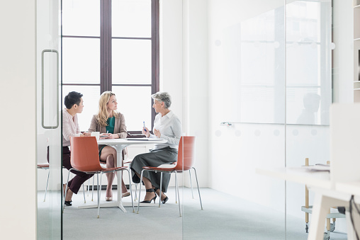 Three businesswomen in business meeting. Three colleagues sitting around a table discussing with window behind.