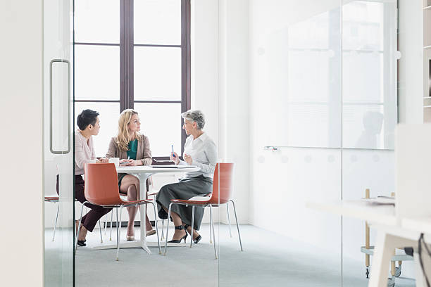 Three women sitting at table in modern office Three businesswomen in business meeting. Three colleagues sitting around a table discussing with window behind. business finance and industry stock pictures, royalty-free photos & images