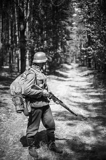 Unidentified re-enactor dressed as German soldier with rifle standing on road in woods. Black and white colors
