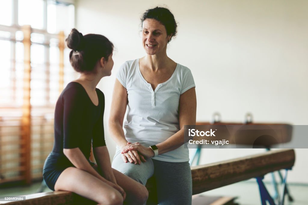 Young Gymnast Girl Talking With Her Coach Cute Little Girl Practicing gymnastics on gymnastics beam. She is sitting on gymnastics beam and talking with her coach of winning on competition. Coach giving her instruction. Exercise in a school gym. She is determined to success. 8-9 Years Stock Photo