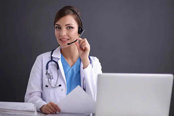 Doctor wearing headset sitting behind a desk with laptop over grey background