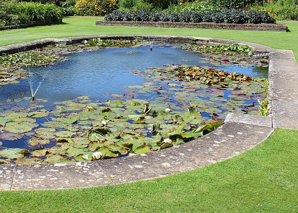 Photo showing an ornamental garden pond with crystal clear water and aeriation provided by an attractive fountain.  Lots of water lily pads provide shade for the goldfish, while the edge of the liner is hidden by paving stones.