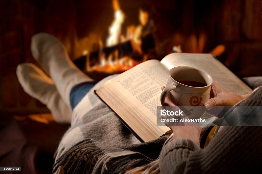 Woman reads book near fireplace Woman resting with cup of hot drink and book near fireplace Drinking Stock Photo