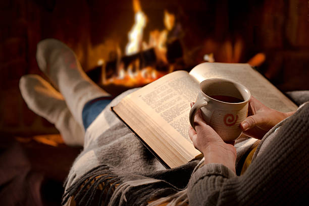 Woman reads book near fireplace Woman resting with cup of hot drink and book near fireplace heat home interior comfortable human foot stock pictures, royalty-free photos & images