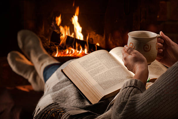 Woman reads book near fireplace Woman resting with cup of hot drink and book near fireplace heat home interior comfortable human foot stock pictures, royalty-free photos & images