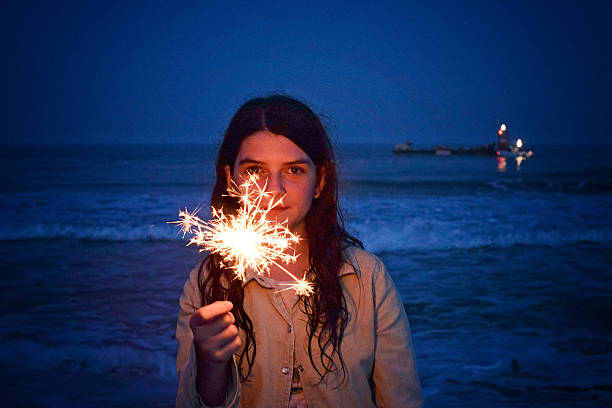 A girl holds a sparkler up by her face on the beach at night.  there is a boat off shore.  The sparkler is sending stars and sparks out from her hand.  She is partially lit from the sparkler.  