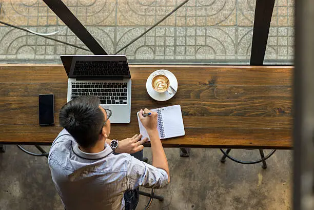 Photo of Young Asian businessman taking note beside laptop on wooden table