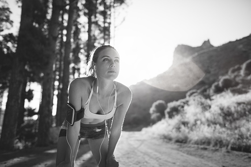 Black and white image of a woman cross-country runner taking a rest with her hands holding onto her knees on a mountain nature trail in early morning sunlight