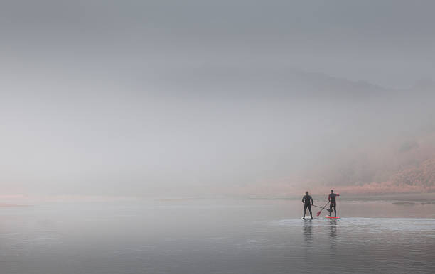 paddle boarding Swansea, UK - April 12, 2016: Two men choose to keep fit by paddleboarding in heavy misty conditions on the river at Three Cliffs Bay, Gower swansea stock pictures, royalty-free photos & images