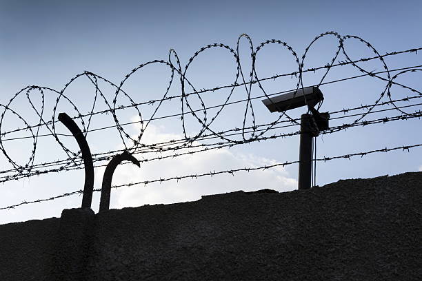 istock Security camera behind barbed wire fence around prison walls 520593276