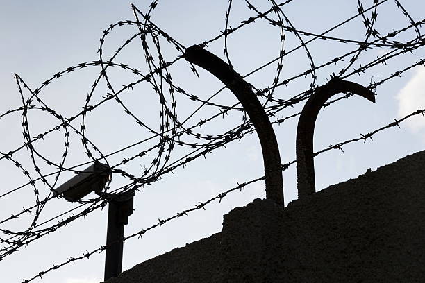 istock Security camera behind barbed wire fence around prison walls 520594168