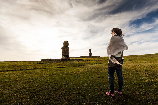 A DSLR photo of a young woman standing in the grass at Ahu Tahai Moai Statue in Easter Island, Chile. She is tucking with a grey scarf. The sun is blocked by some clouds, so the entire scene is lit by a very soft light.