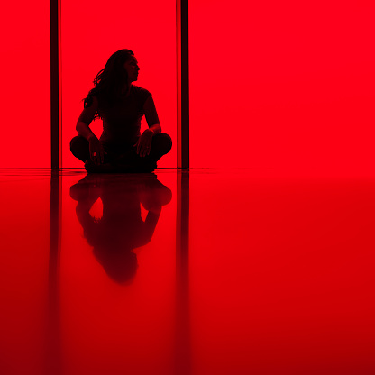 silhouette of a young woman sitting cross-leg against a red window background.