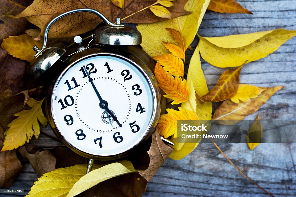 Retro clock on a wooden table with autumn leaves Retro metal clock standing on wooden table with leaves laying around Alarm Clock Stock Photo