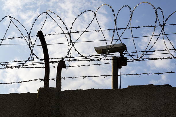 istock Security camera behind barbed wire fence around prison walls 520608286