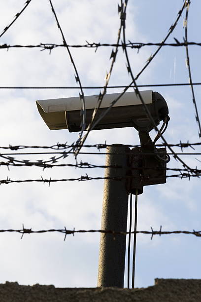 istock Security camera behind barbed wire fence around prison walls 520609478