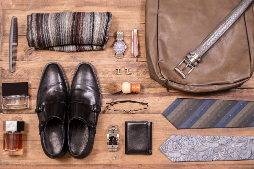 Variety of men's accessories organized in knolling arrangement.  Items include: shoes, tie, glasses, scarf, wallet, watch, shaving supplies, cologne, belt, knife.  The items all lie on a wooden desk or table.   Men's personal accessories, clothing themes.  Father's Day.  Fashion.  Business.  Retail.