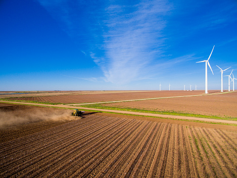 A tractor is plowing up rows of barren land for future crops. The dirt is dry and dusty. Dust is kicked up by the tractor. A windmill farm is in the distance with rows of white windmills. Dry land is seen up to the horizon. The sky is bright blue with spotty clouds.