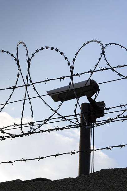 istock Security camera behind barbed wire fence around prison walls 520612264