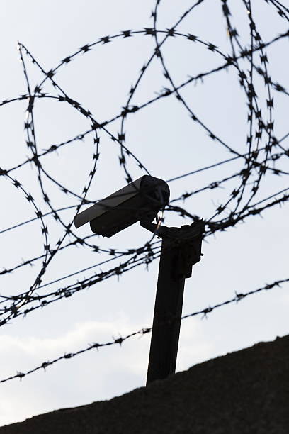 istock Security camera behind barbed wire fence around prison walls 520612400