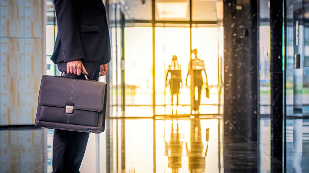 Businessman with suitcase waiting in the lobby Side view of a businessman with a suitcase waiting in a lobby. Two figures approaching in the background. walking into glass door stock pictures, royalty-free photos & images