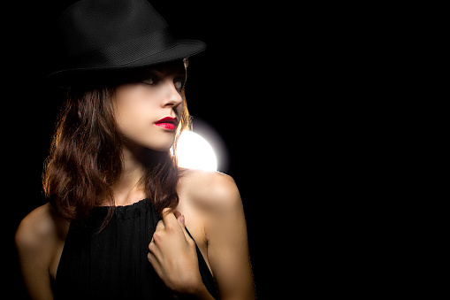 Actress with classic smoky dark make up in Hollywood film noir style.  She is wearing a black noir style fedora hat.  She is also lit in directional light source indicative of the noir style. The image depicts the trend of dark noir style make up in cosmetic fashion.