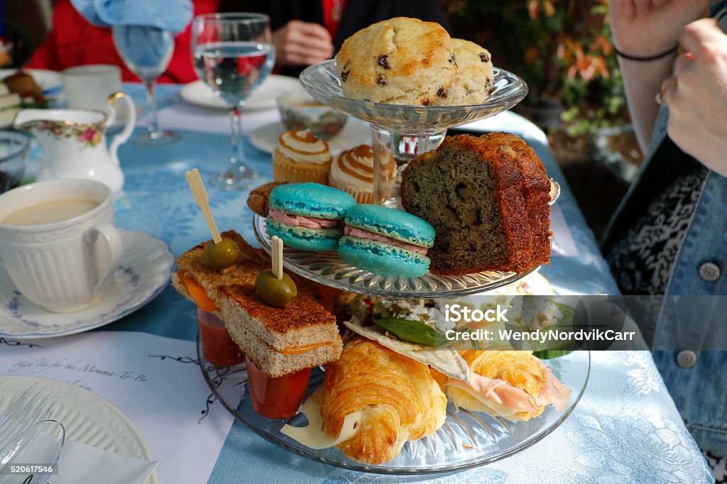 Traditional high tea baby shower served in the garden Traditional luxurious English high tea to celebrate a baby shower with family and friends for the mother to be in a Vancouver garden on a fine spring day. A three-tiered tray of delicate finger sandwiches and baked goods of savory  and sweet selections including: grilled cheese with chilled tomato soup, French macarons, salted caramel mini cupcakes, banana loaf, raisin scones, ham and cheese croissants. Served on a blue linen tablecloth with fine china.  Afternoon Tea Stock Photo