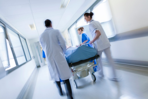 A motion blurred photograph of a patient on stretcher or gurney being pushed at speed through a hospital corridor by doctors & nurses to an emergency room