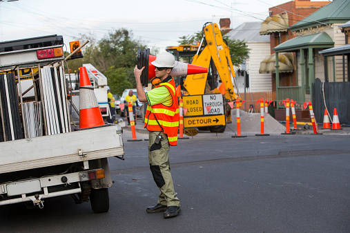 A young traffic controller worker in his twenties wearing dark safety glasses, a white hard hat and a high visibility orange vest is working on the side of the road packing large traffic orange cones back in the tray of a white utility truck. The road is closed with orange poles in front of an yellow excavator parked next to a pile of gravel. The day is overcast, the shy is white and there is no shadows. It is a suburban street with old weatherboard houses