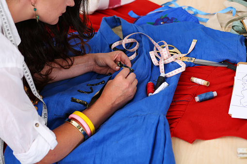 Female fashion designer sewing accessories to blue retro style dress with needle and threads. Creating garment, clothes sewing and repair service, seamstress at work in workshop concept