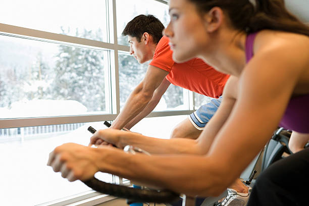 Man and woman using exercise bikes in gym, looking through window at snowy scene, side view