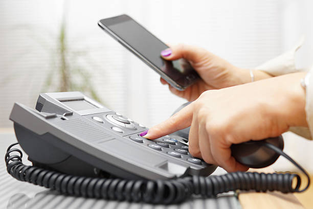 Woman is dialing on land line phone and looking on mobile phone stock photo