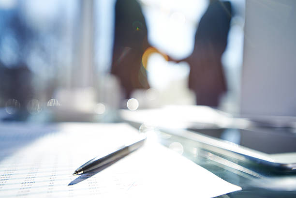 Blurred business success Document and pen on the desk, silhouettes of business people shaking hands in the background financial report photos stock pictures, royalty-free photos & images
