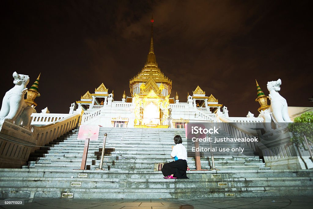 Praying Thai woman at Wat Traimit in night Bangkok, Thailand - October 29, 2014: Night shot of an adult thai woman sitting and praying on steps of temple Wat Traimit in Chinatown. On top of steps is an image of king Bhumipol. At both sides of steps are lion sculptures. Adult Stock Photo